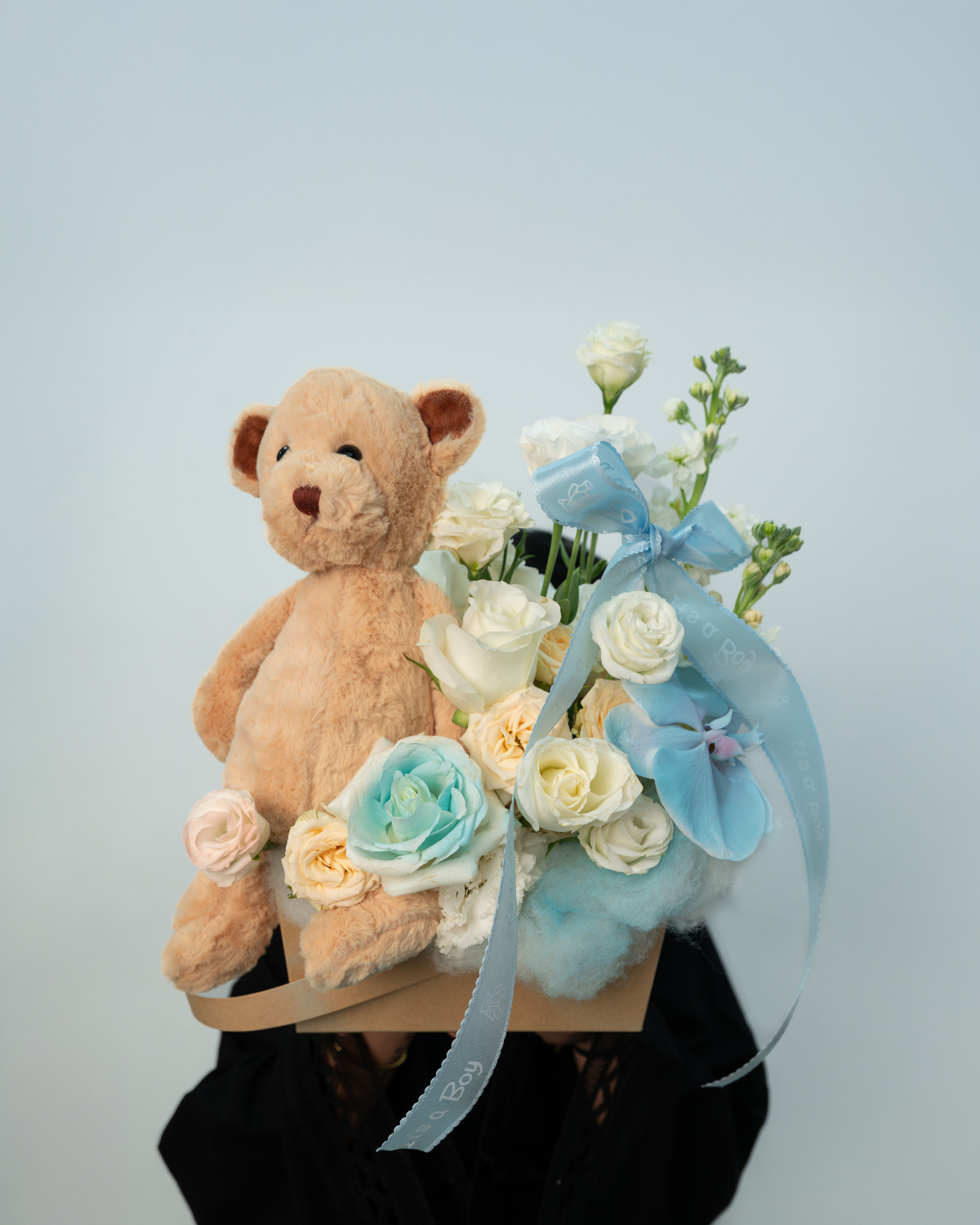 A floral arrangement with a cut phalaenopsis orchid, white roses painted blue, lisianthus, and a teddy bear, all arranged in a rustic kraft box.