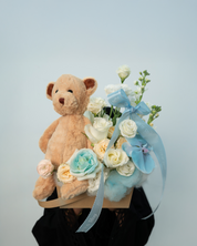 A floral arrangement with a cut phalaenopsis orchid, white roses painted blue, lisianthus, and a teddy bear, all arranged in a rustic kraft box.